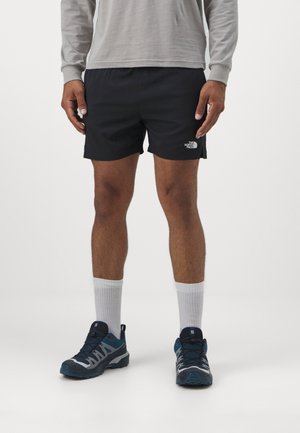 Mężczyzna ubrany w czarne spodenki The North Face, szarą bluzę z długim rękawem, białe skarpetki i niebiesko-szare buty sportowe, stojący na jednolitym tle.