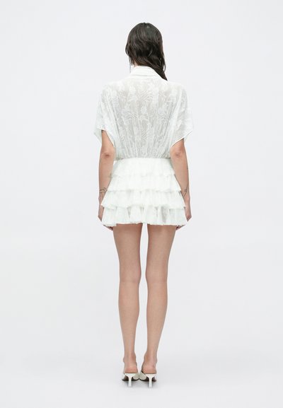 Biele kvetinové krajkové mini šaty s krátkymi rukávmi a viacvrstvovou volánovou sukňou. Tkanina je priehľadná, čím zvýrazňuje jemnú textúru a vzor.
