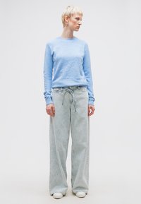 Osoba o krótkich blond włosach, ubrana w jasnoniebieski sweter, szerokie dżinsy z jasnego jeansu z paskiem do wiązania oraz białe trampki, stojąca i patrząca w prawo.