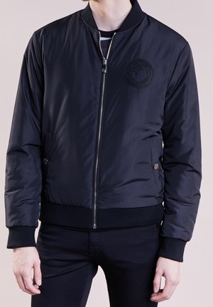 Man draagt een zwart bomberjack met ritssluiting, geribde manchetten en tailleband, geperst logo op de borst, en handen ontspannen langs het lichaam.