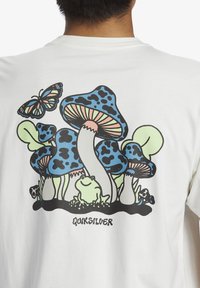 White t-shirt featuring a colorful graphic of blue-speckled mushrooms, a green frog, and a butterfly, with "Quiksilver" text at the bottom.