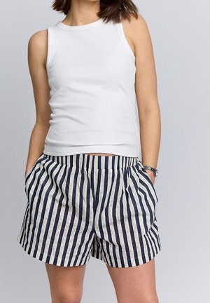 Personne portant un top blanc sans manches en côte et un short à rayures verticales bleu marine et blanc taille haute, les mains dans les poches.