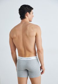 Sive pamučne bokserice s bijelim pojasom na kojem je natpis "Calvin Klein"; prikazane s leđa na muškom modelu.