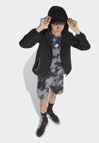 Čierny vetrovka s kapucňou, šedé športové tričko s vzorom, zodpovedajúce šortky, čierne ponožky a tenisky. Obsahuje zipsové vrecká a logové akcenty.