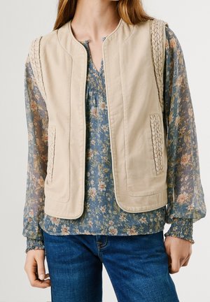 Beige mouwloos vest met gevlochten rand, gedragen over een blauwe bloemenlange mouwen blouse en een blauwe spijkerbroek bij een staand persoon.