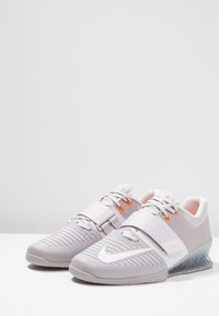 Grå atletiske sko med en tekstureret overdel, der har en midt-fodstrop og orange accenter. Gennemsigtig sål med et futuristisk design.