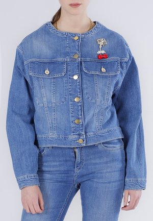 Donna che indossa una giacca di jeans blu con bottoni e tasche sul petto, con una spilla a forma di ciliegia sul lato sinistro, abbinata a jeans di jeans blu.