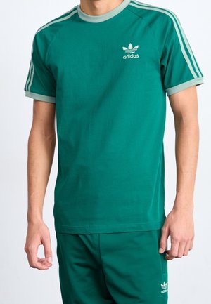 Osoba nosząca zieloną koszulkę Adidas i pasujące spodnie, oba z jasnooliwkowymi akcentami i logo Adidas na piersi i nodze.