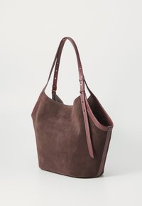 Borsa tote in suede marrone con manici in pelle scura regolabili e dettagli in oro, mostrata in piedi su uno sfondo neutro.