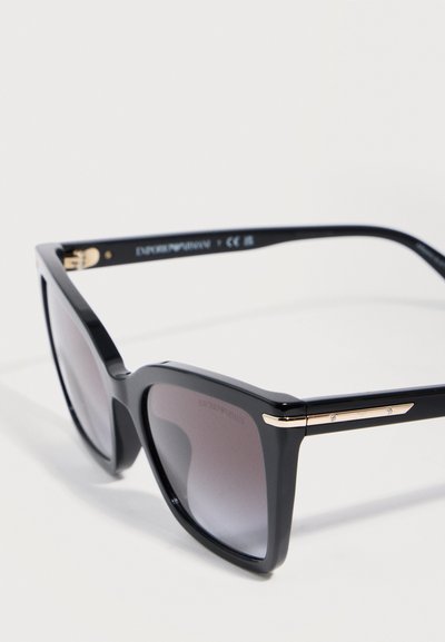 Μαύρα γυαλιά ηλίου με τετράγωνο πλαίσιο, φακούς με βαθμιαία σκουρότητα και χρυσές λεπτομέρειες στις βραχίονες, ακουμπισμένα σε λευκή επιφάνεια.
