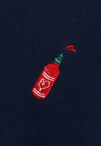 Tissu bleu marine présentant une petite bouteille de ketchup rouge brodée avec un bouchon vert et des accents blancs, arborant un design en forme de cœur sur la bouteille.