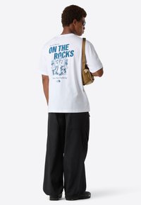 Person med hvid grafisk t-shirt med teksten "ON THE ROCKS" og tegneserie-terninger, sorte bukser, solbriller og bærer en beige skuldertaske.