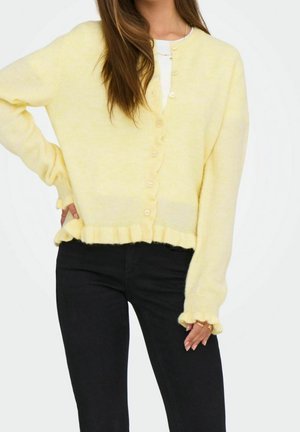 Woman wearing soft yellow buttoned cardigan with ruffled edges and black pants, hand resting on hip.