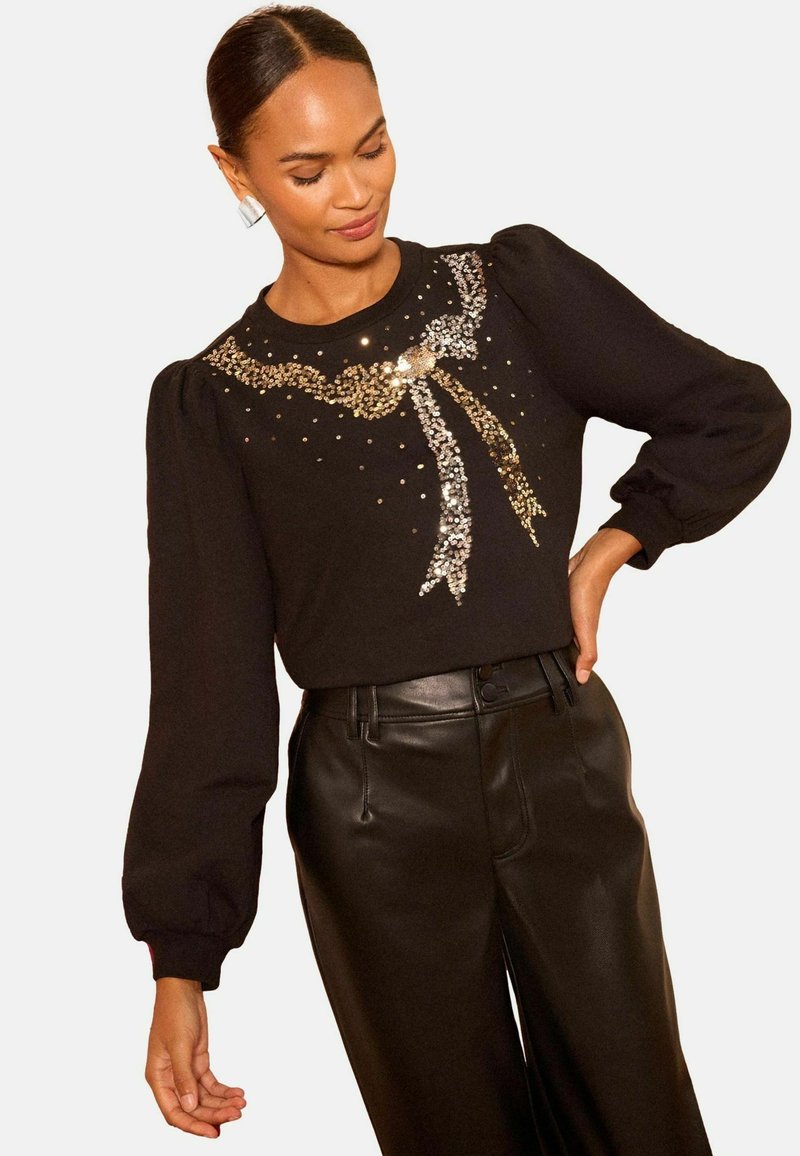 Suéter negro con mangas abullonadas que presenta un diseño de lazo metálico hecho de lentejuelas doradas y plateadas. Combinado con pantalones negros de talle alto.