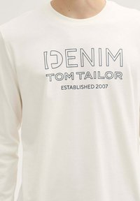 Fehér, hosszú ujjú póló, amelyen sötétkék betűkkel a "DENIM TOM TAILOR ESTABLISHED 2007" felirat található. Pamut anyag, kerek nyakkivágással.
