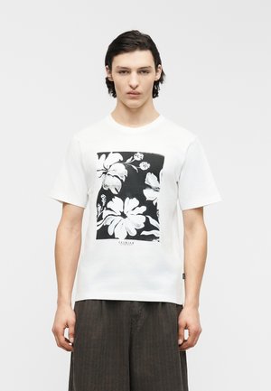 Noor mees tume juustega, kandes valget lühikese varrukaga t-särki, millel on must ruut valgete lillemotiividega, koos tumedate pükstega.