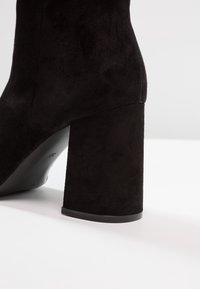 Botín negro de ante hasta el tobillo con tacón cuadrado, superficie texturizada y suela de goma negra lisa. Presenta un diseño minimalista y costuras sutiles.