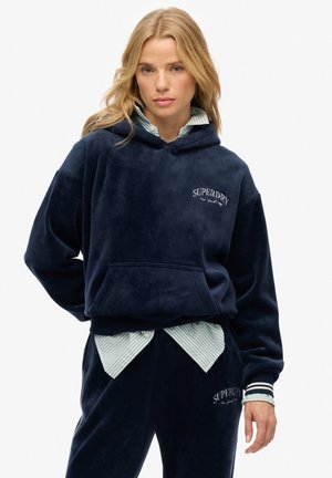 Kvinde med blondt hår bærer en mørkeblå ribstrikket hættejakke og matchende bukser med en hvidstribet skjorte under. Både hættejakken og bukserne har Superdry-logoet.
