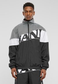 Black zip-up jacket with mesh paneling at the top. Features white horizontal stripe with bold black text. Elastic cuffs and hem.