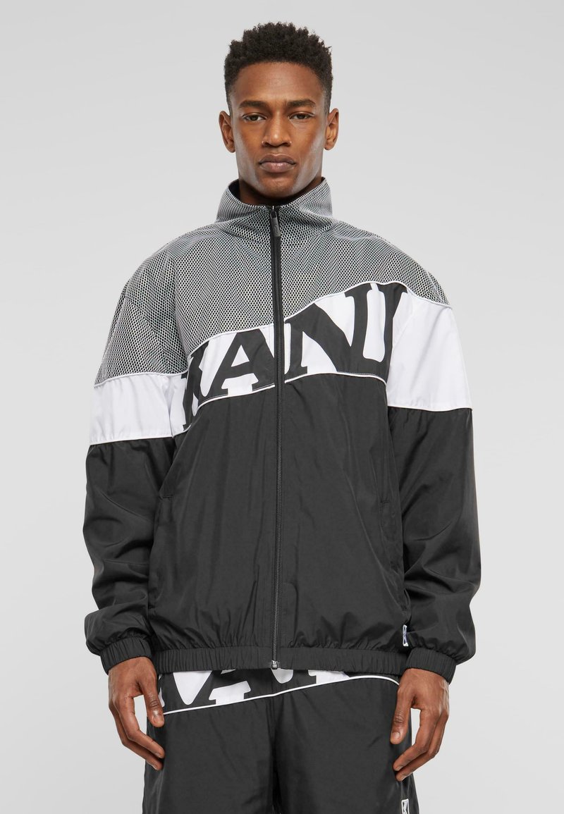 Black zip-up jacket with mesh paneling at the top. Features white horizontal stripe with bold black text. Elastic cuffs and hem.