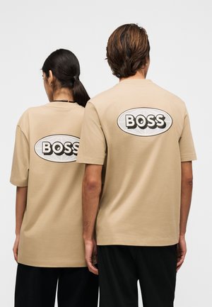 To personer står rygg mot rygg iført beige t-skjorter med en stor hvit oval logo og ordet "BOSS" i fete svarte bokstaver på baksiden.