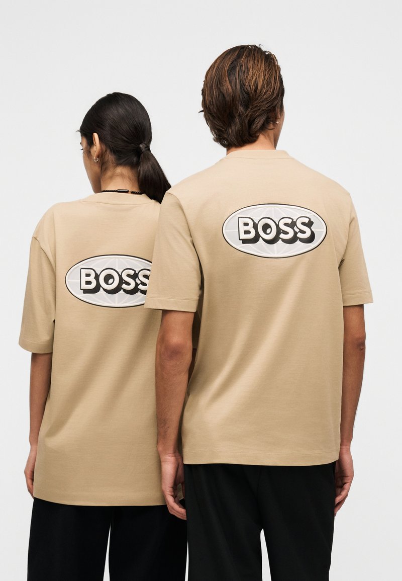 Deux personnes se tiennent dos à dos portant des t-shirts beige avec un grand logo ovale blanc et le mot « BOSS » en lettres noires en gras dans le dos.