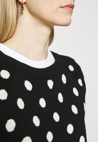 Schwarzer Pullover mit großen weißen Punkten, gestrickter Textur und Rundhals-Ausschnitt. Getragen über einem weißen T-Shirt mit Rundhalsausschnitt.