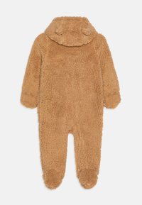 Brązowy, puszysty kombinezon wykonany z miękkiego, teksturowanego materiału, z kapturem z małymi uszami i zakrytymi stopami. Widok od tyłu.