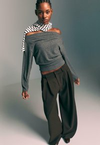 Szary sweter typu off-the-shoulder z prążkowanym dołem, zestawiony z szerokimi brązowymi spodniami. Czarne botki i paskowany szalik dopełniają całość.