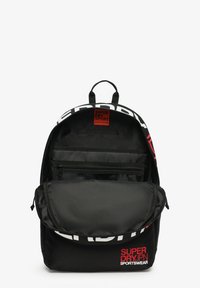 Zaino sportivo Superdry nero con scomparto principale aperto che mostra tasche interne con cerniera e in rete, branding bianco e rosso sul davanti.