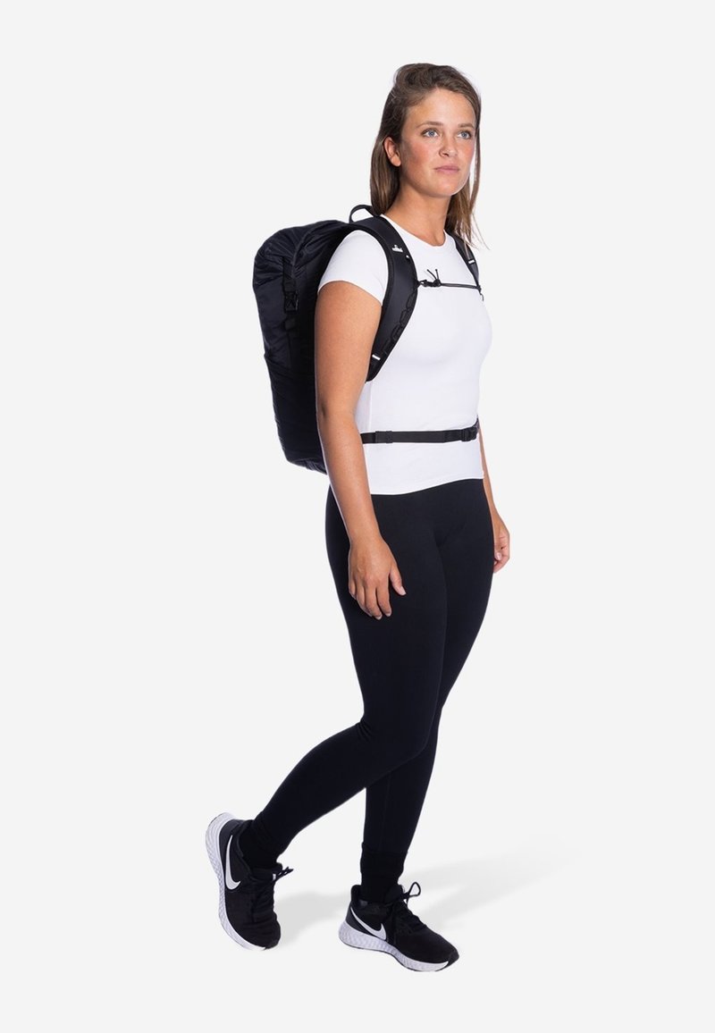 Zwart rugzak met verstelbare schouderbanden, gedragen over een witte top en zwarte leggings. Gladde stof met minimale accenten. Neutrale achtergrond.