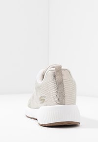 Zapatilla con un upper de un dorado metálico, diseño texturizado, media suela blanca y suela de goma marrón. Incluye un tirador en el talón.