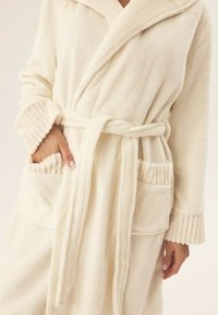 Kremowy polarowy szlafrok z kołnierzem szalowym, paskiem w talii, prążkowanymi mankietami i dwiema przednimi kieszeniami. Miękka tekstura i luźny krój.