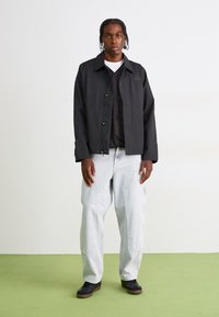 Veste légère noire avec un col, fermeture à boutons et poches latérales, superposée à un gilet matelassé noir, associée à un jean baggy gris clair et des chaussures noires.