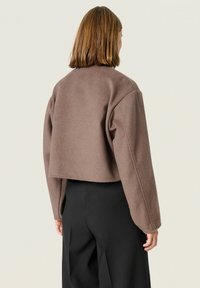 Brun cropped jakke lavet af blødt stof med sænkede skuldre og brede ærmer. Båret over sorte højtaljede bukser.
