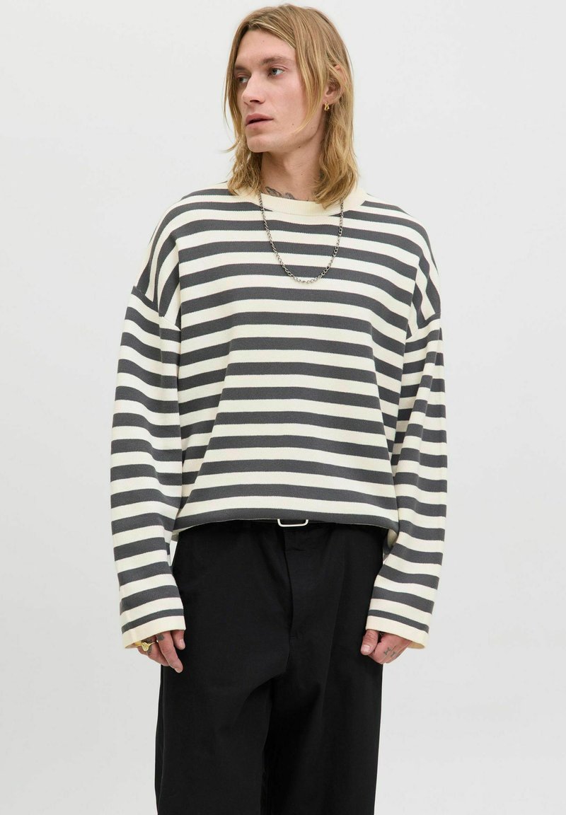 Striped sweater in cream and dark gray. Relaxed fit with long sleeves, dropped shoulders, and a rounded neckline. Paired with black trousers.