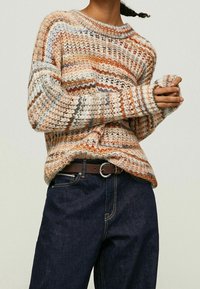 Femme portant un pull en tricot épais multicolore et un jean bleu foncé avec une ceinture marron, les mains jointes devant elle.