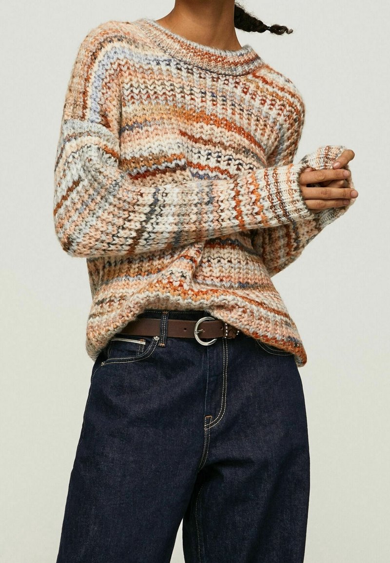 Femme portant un pull en tricot épais multicolore et un jean bleu foncé avec une ceinture marron, les mains jointes devant elle.