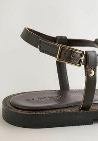 Gros plan d'une sandale en cuir brun foncé avec boucle dorée, semelle plate et coutures visibles sur les brides et la semelle intérieure portant la mention "NEXT."