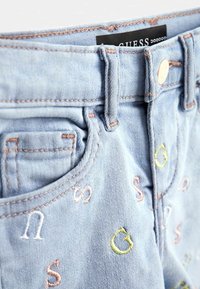 Jasnoniebieskie dżinsy z różowymi i żółtymi haftowanymi literami rozrzuconymi po tkaninie w pobliżu przedniej kieszeni i pasa.