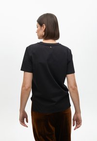 T-shirt nera in cotone con maniche corte e scollatura rotonda. Presenta un piccolo logo dorato sul retro. Abbinato a pantaloni in velluto marroni.