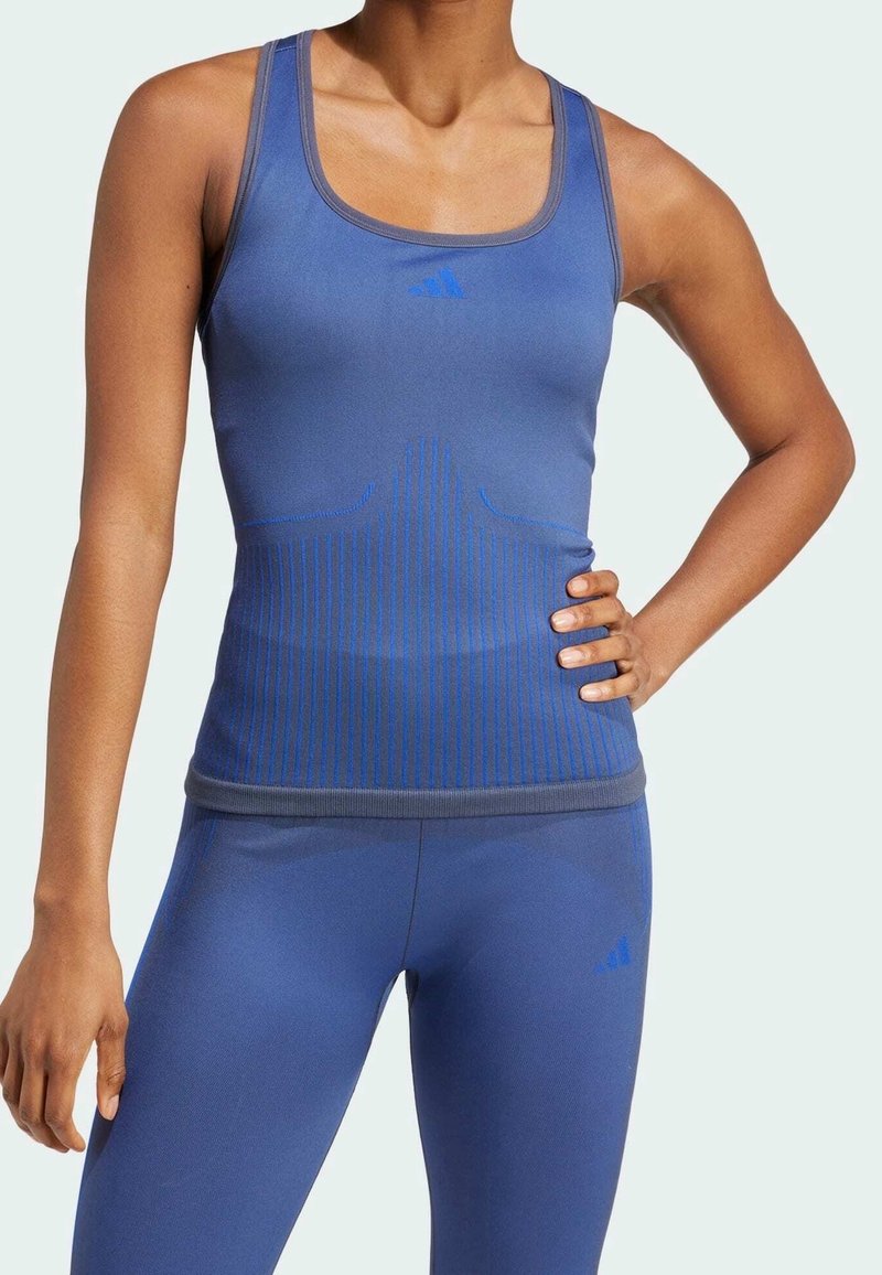 Modrý atletický top s přiléhavým střihem, vyrobený z hladkého materiálu, se svislými pruhy a kontrastním lemem okolo výstřihu.