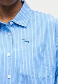 Mėlyna ir balta vertikaliomis juostelėmis dryžuota marškiniai su sagomis, apykakle, krūtinės kišenė ir siuvinėta užrašu „Obey“ virš kišenės.