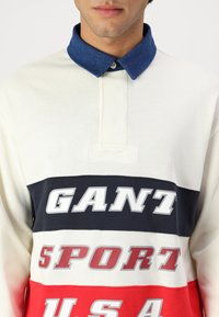 Polo skjorte med krage i blå denim; hvite og fargede striper med teksten "GANT SPORT USA" i fet skrift. Laget av bomull, med avslappet passform.