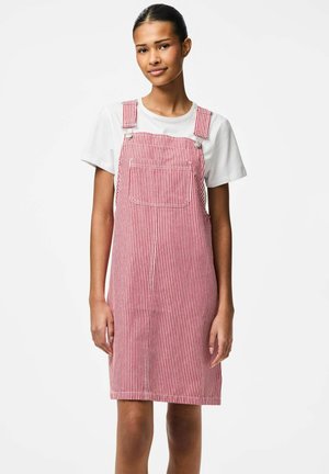 Kobieta nosząca białą koszulkę pod czerwoną i białą, pionowo paskowaną sukienkę bez rękawów z przednią kieszenią.