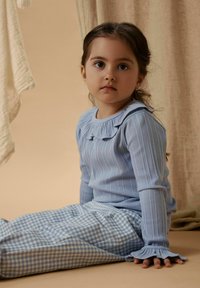 Jasnoniebieska prążkowana bluzka z długim rękawem i falbanianym kołnierzem, w połączeniu z niebiesko-białymi spodnimi w kratę. Miękki, teksturowany materiał, casualowy krój.