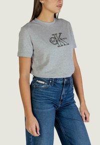 Szary bawełniany T-shirt z czarnym logo "Calvin Klein". W połączeniu z jeansami o wysokim stanie w ciemnym niebieskim kolorze. Krótkie rękawy, klasyczny krój.