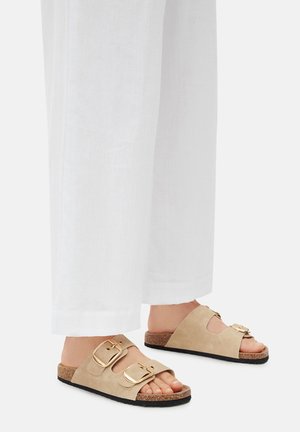 Sandalias de ante beige con dos hebillas doradas, plantilla de corcho y suela exterior de goma negra. Se llevan con pantalones de lino blancos.