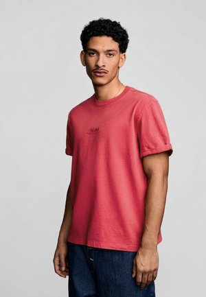 Man with short curly hair wearing a red short-sleeve t-shirt and blue jeans, standing against a plain light gray background.