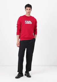 Sudadera roja con textura acanalada y logo blanco "KARL LAGERFELD", combinada con pantalones negros y zapatos negros. Diseño simple y casual.